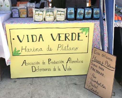 A table at a market with plantain flour and coffee for sale; the sign reads: "Vida Verde, Harina de Platano", Asociación de Producción Alimenticia Defensoras de la Vida