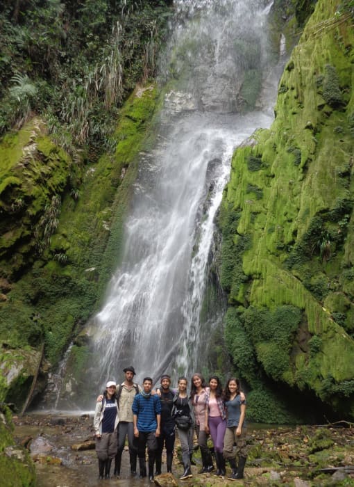 group of people stands before a waterfall that rushes down a mossy green rock face located in the Intag Valley